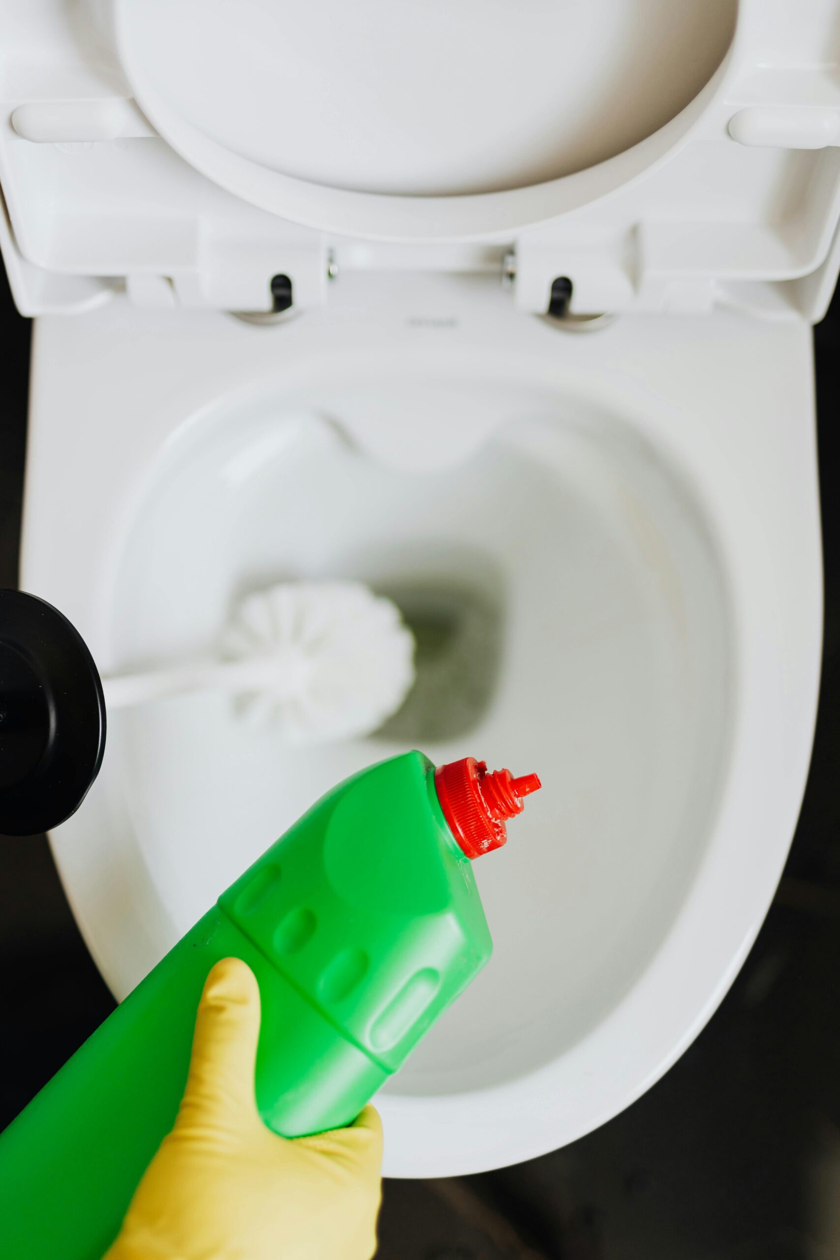 a hand wearing a yellow glove holds a bottle of cleaning bleach as they clean a toilet
